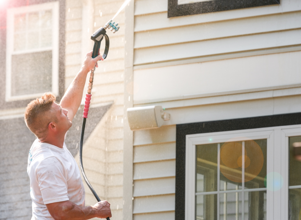 A person power-washes the exterior of a house, aiming the spray upward, with sunlight creating lens flares on the window.