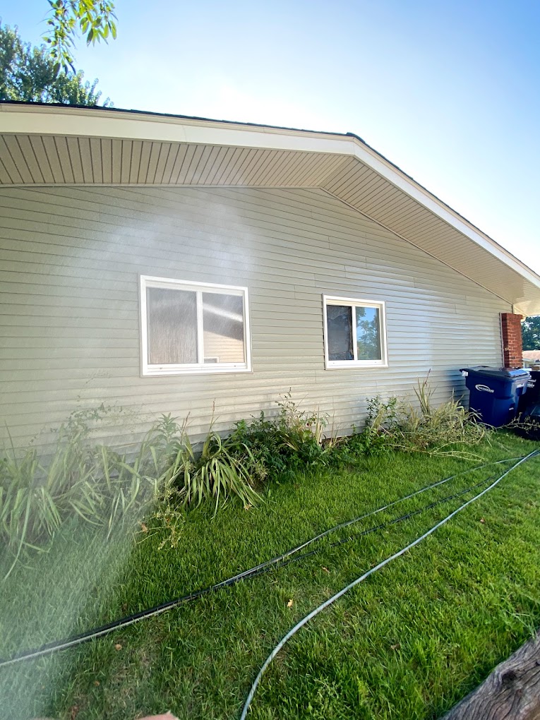 A gray house with white windows, surrounded by plants, green lawn, hose, and blue recycling bins, is misted. Clear sky above.