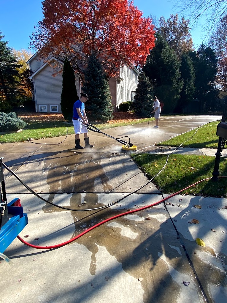 Two people pressure-wash a driveway on a sunny day. Vibrant autumn foliage and a suburban house create a colorful, serene background.