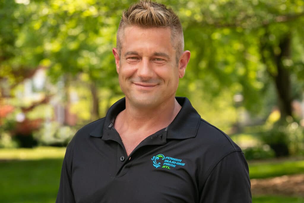 A person with short hair smiles outdoors in a park. Wearing a black polo with a blue logo, surrounded by green foliage.