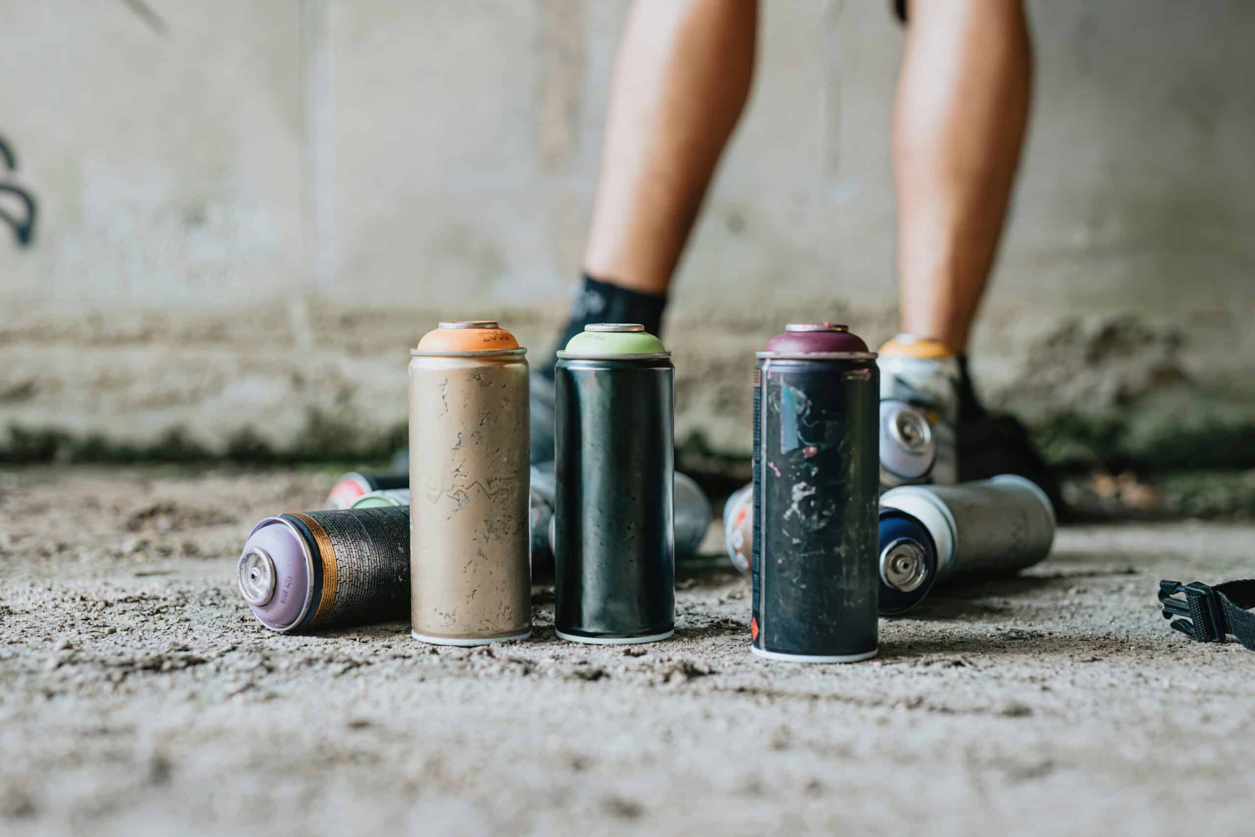 Spray paint cans are scattered in front of a person's legs, standing on a rough concrete surface in an urban setting.