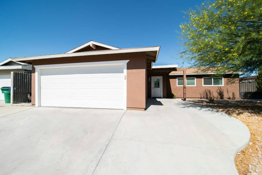 Single-story house with a brown exterior, white garage door, and concrete driveway, set against a clear blue sky with a tree nearby.