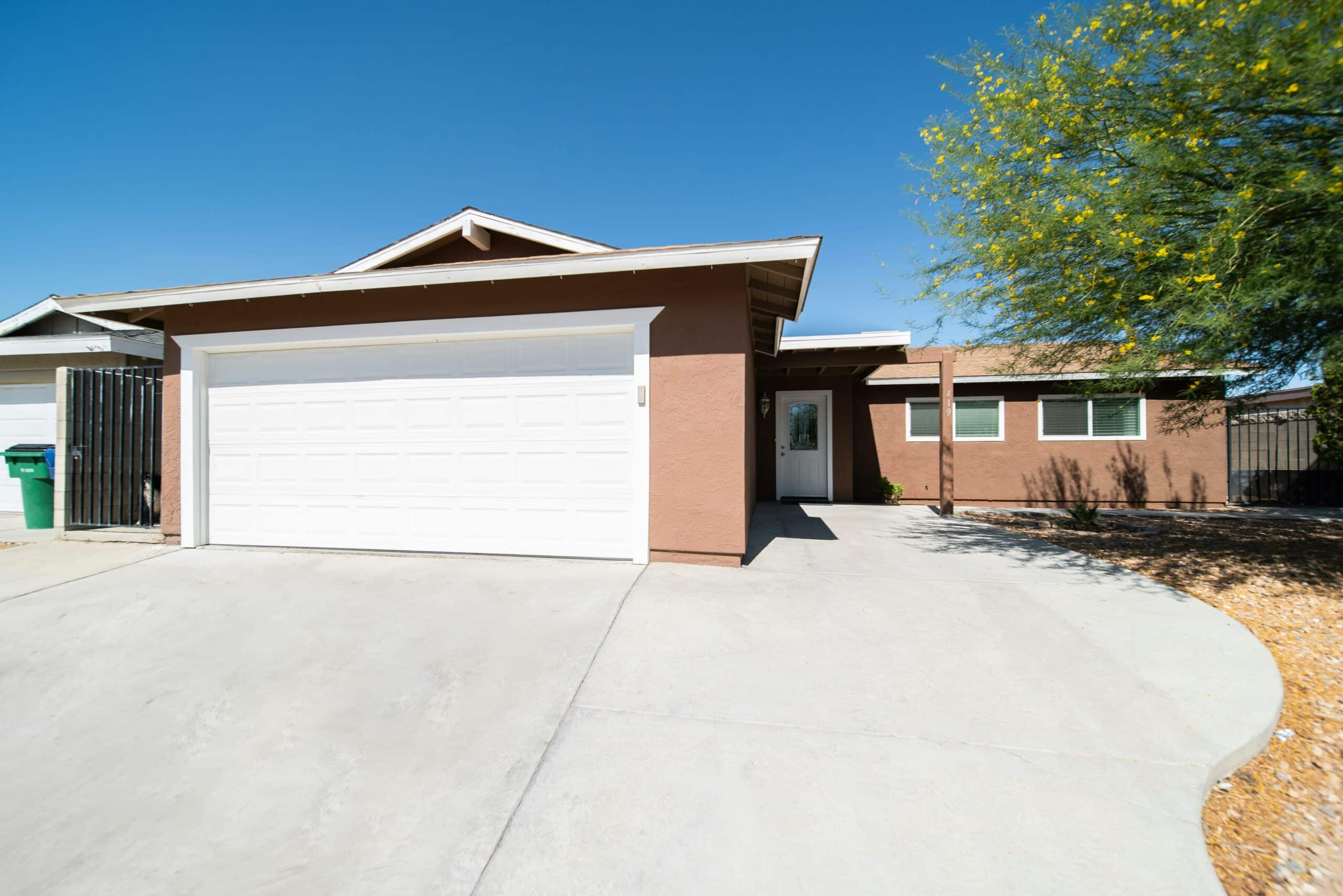Single-story house with a brown exterior, white garage door, and concrete driveway, set against a clear blue sky with a tree nearby.