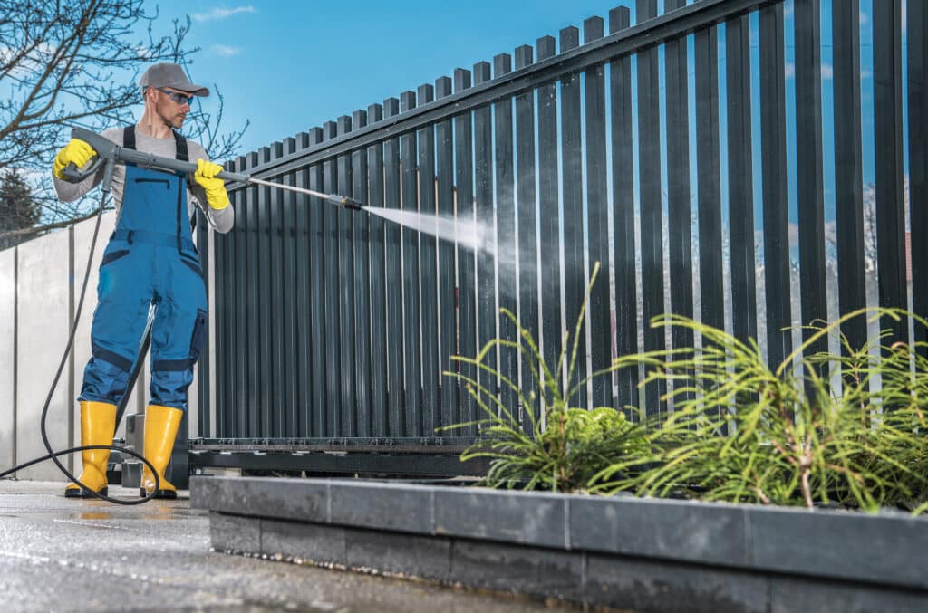 A person in protective gear pressure washes a metal fence near green plants under a clear blue sky.