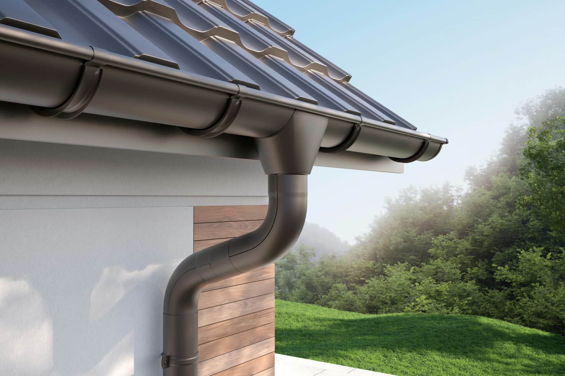 A close-up of a house roof with metal gutters and downspout, surrounded by lush greenery under a clear blue sky.