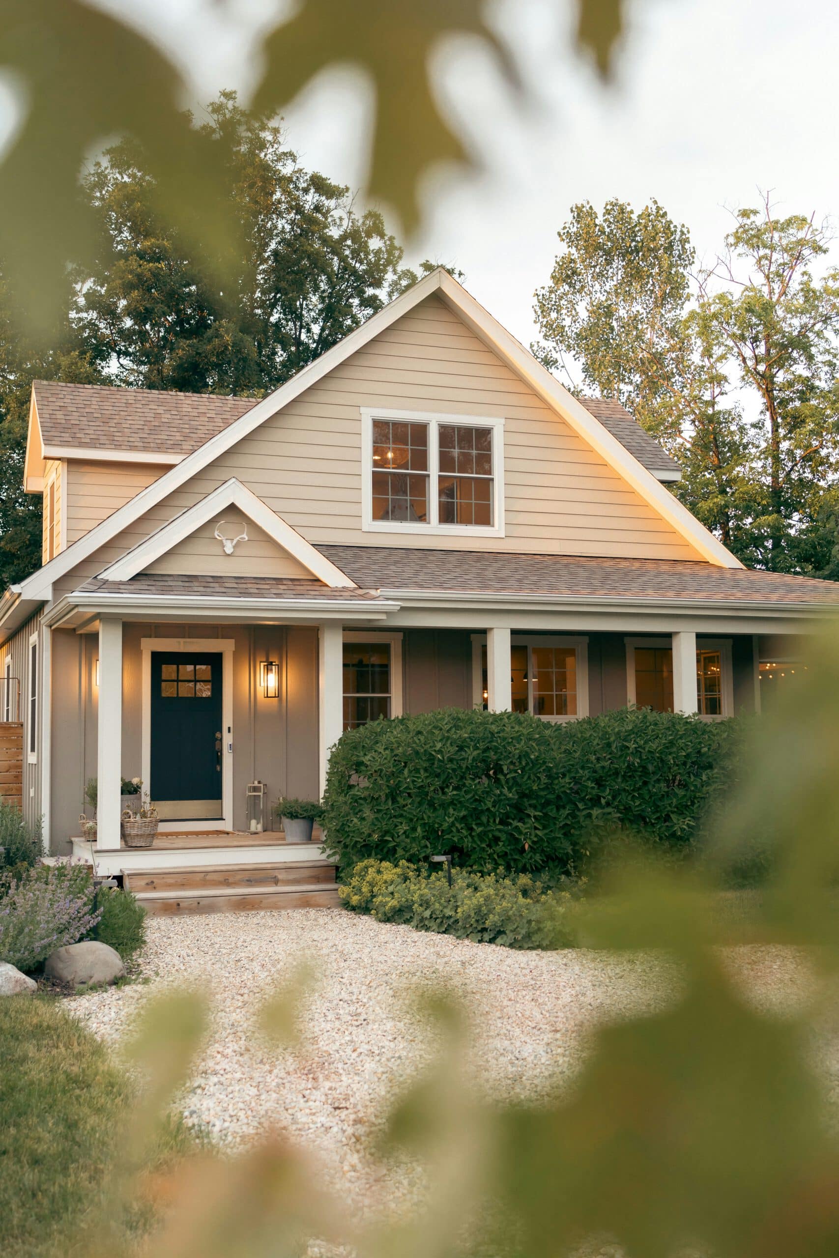Charming house with a cozy porch, surrounded by lush greenery and a gravel path, set against a backdrop of tall trees.