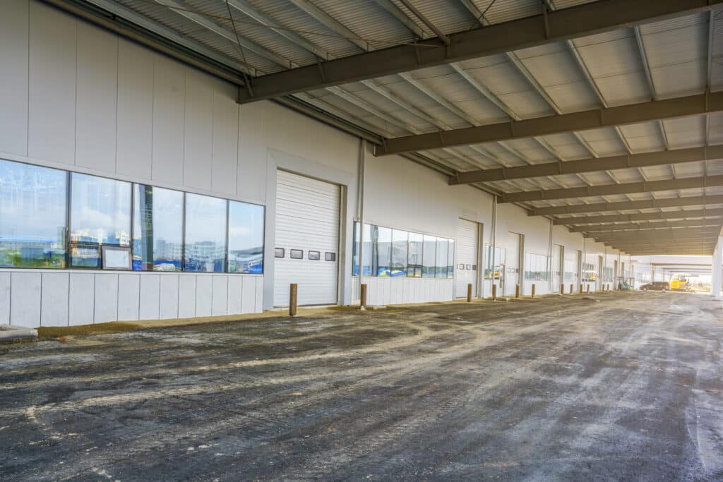 Large industrial warehouse interior with multiple garage doors, high ceiling, and windows reflecting outside construction. No people or landmarks visible. Empty asphalt floor.