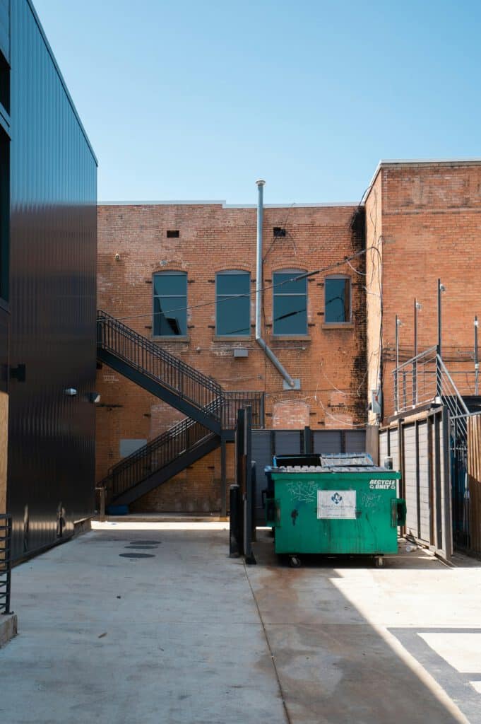 Brick building with fire escape stairs, green recycling dumpster in alley, clear blue sky above, industrial setting, empty and quiet atmosphere, urban environment.