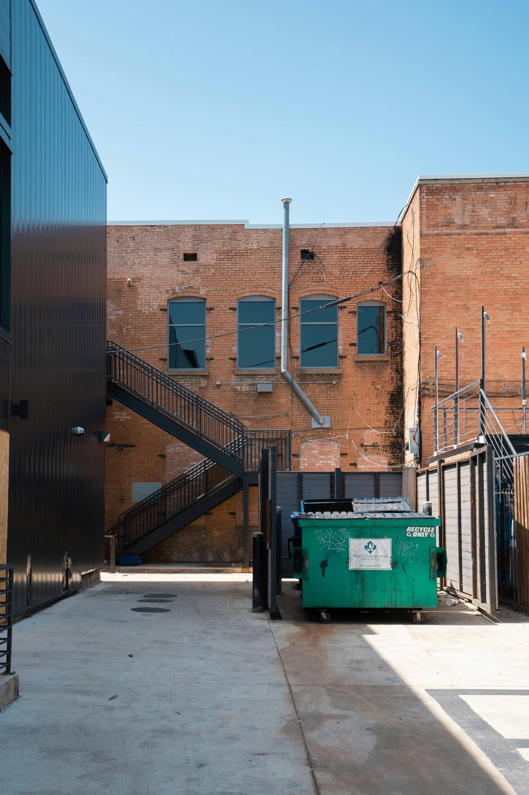 Brick building with fire escape stairs, green recycling dumpster in alley, clear blue sky above, industrial setting, empty and quiet atmosphere, urban environment.