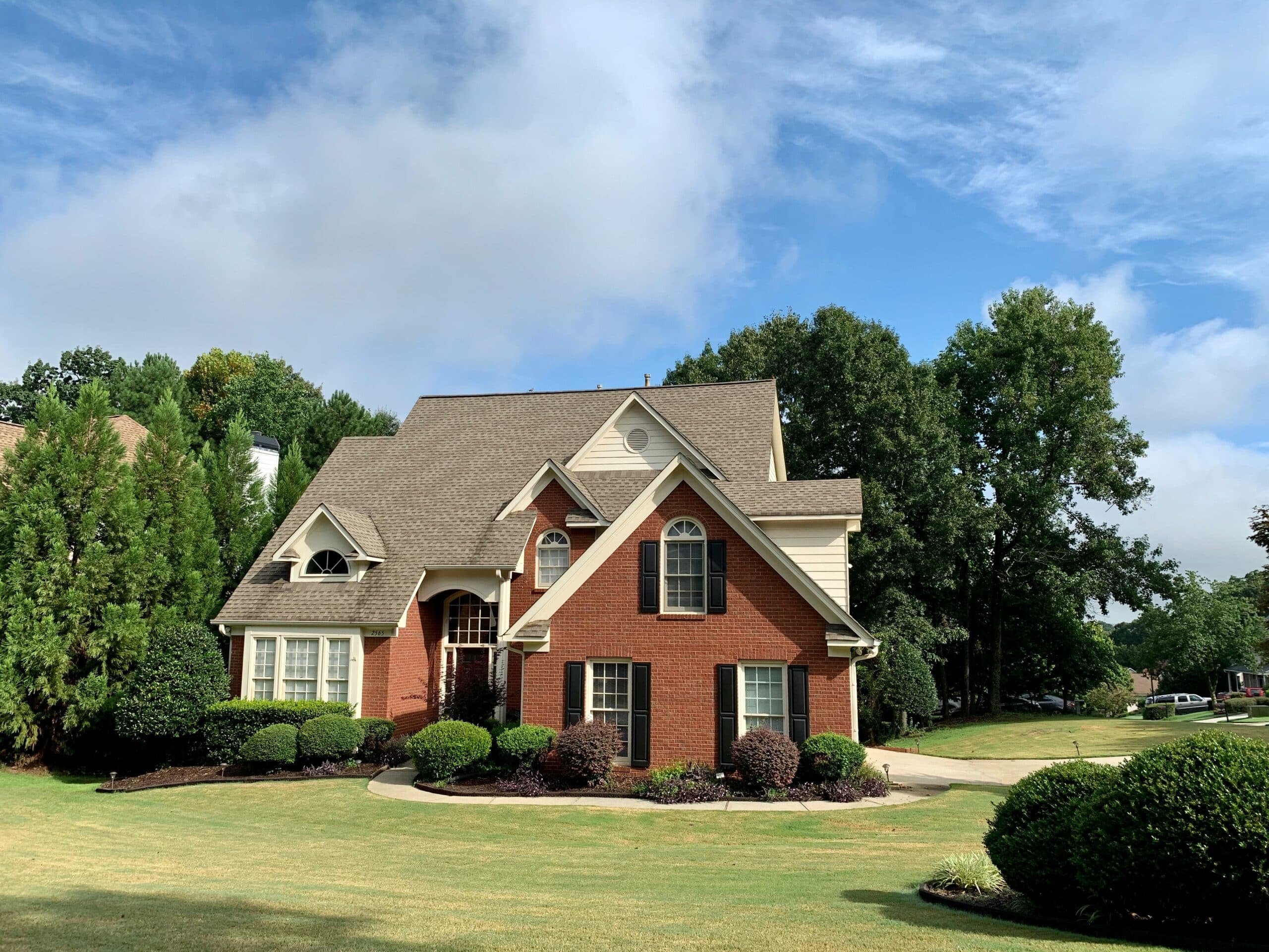 A charming two-story red brick house with manicured lawn, surrounded by lush green trees under a blue sky with clouds.