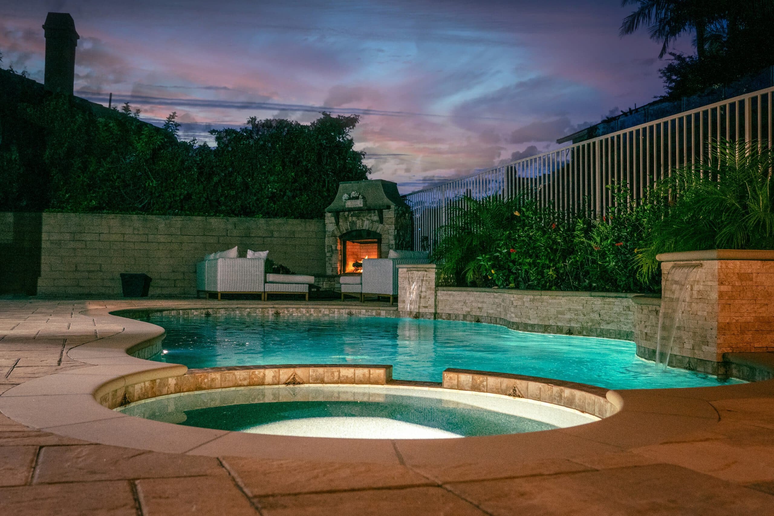 Outdoor pool and jacuzzi with modern brick design at twilight, surrounded by lush greenery and patio furniture, creating a serene, inviting atmosphere.