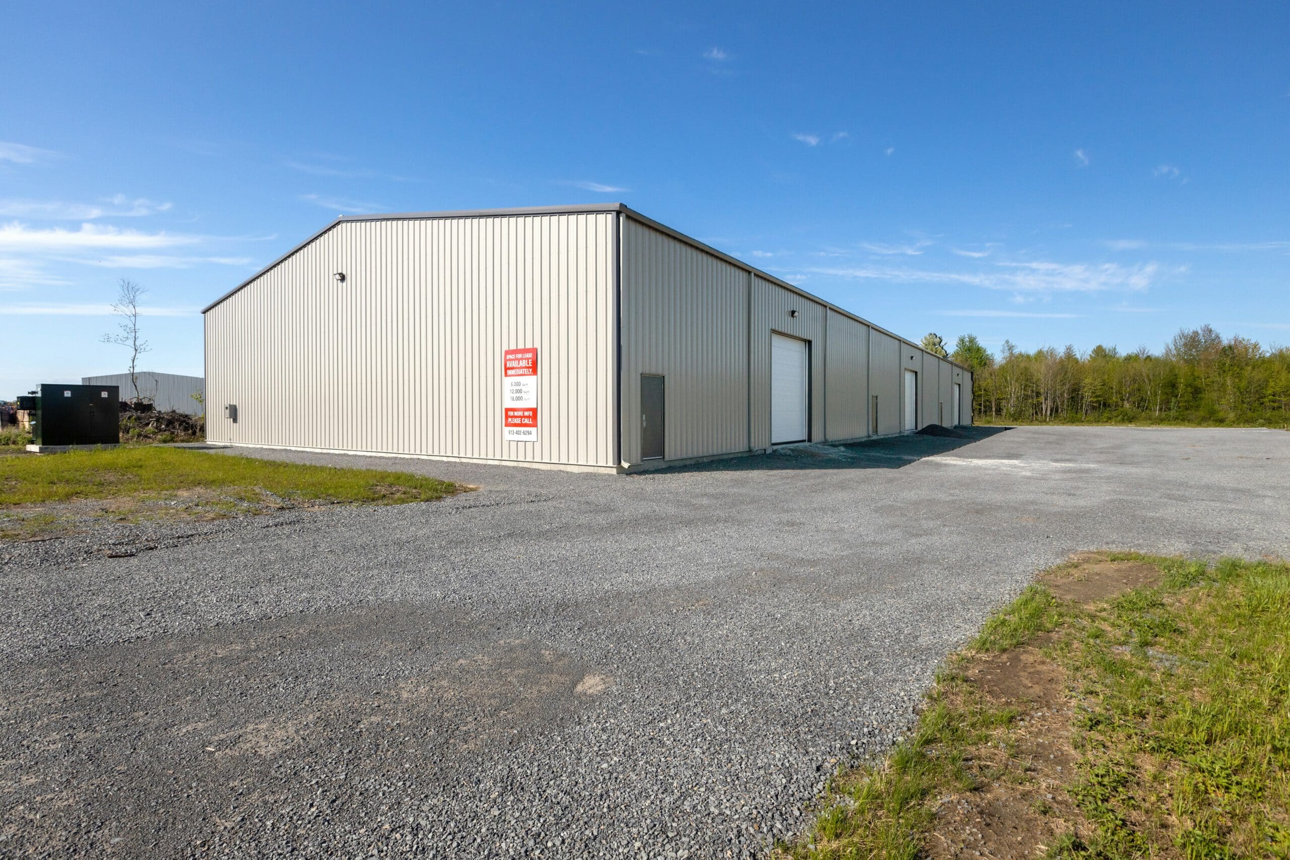 A large, beige industrial warehouse sits on a gravel lot surrounded by trees under a clear blue sky, featuring a sign for lease.