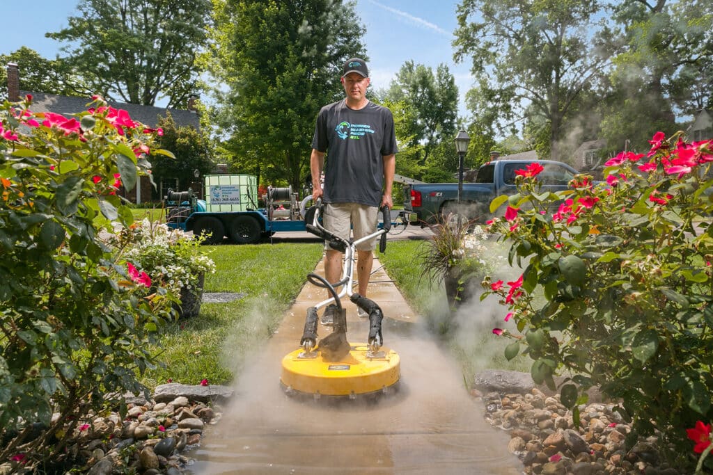 A person uses a yellow pressure washer on a garden pathway, surrounded by pink flowers and greenery, with a truck and equipment nearby.