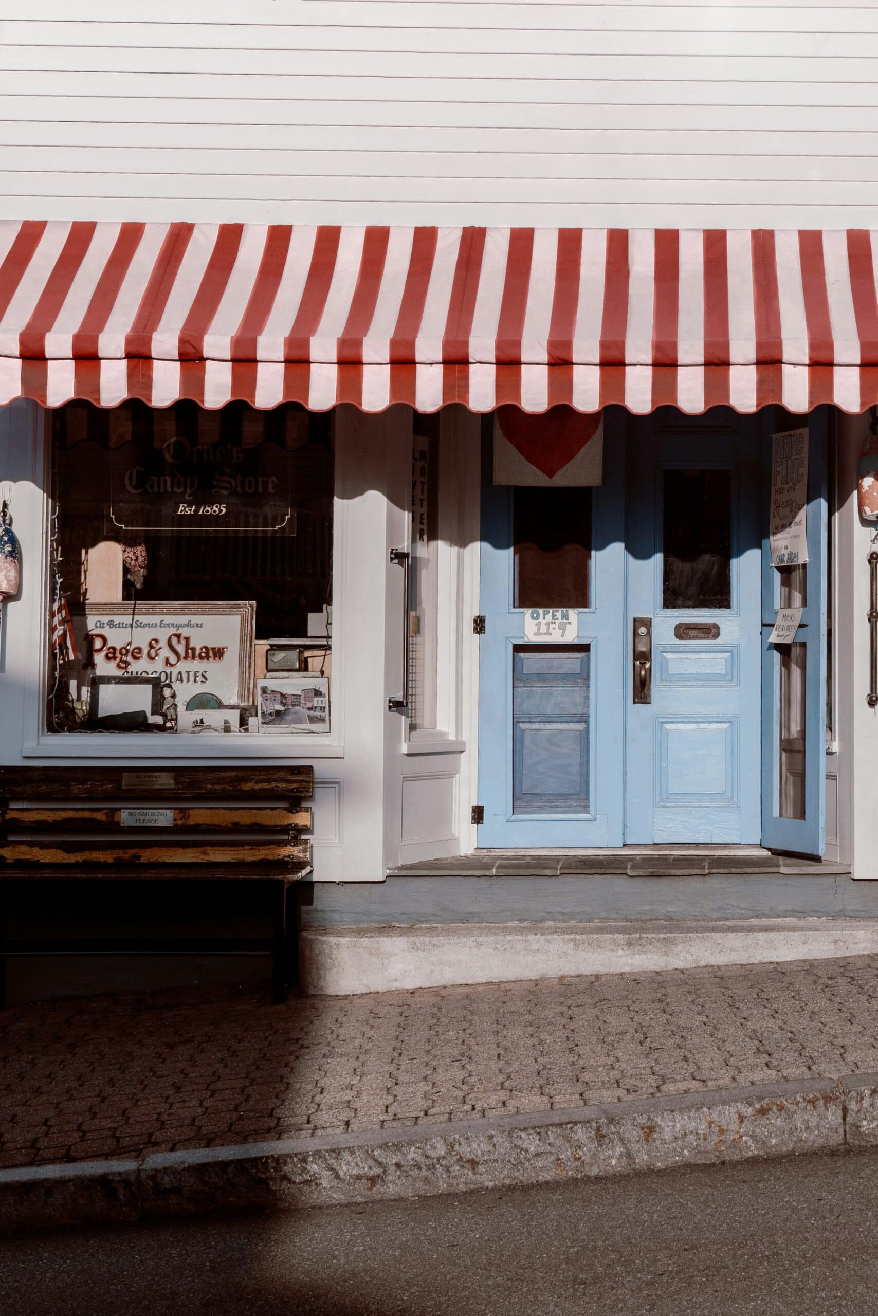 Vintage candy store with blue doors and striped red awning, featuring a welcoming bench and nostalgic signage under sunny skies.