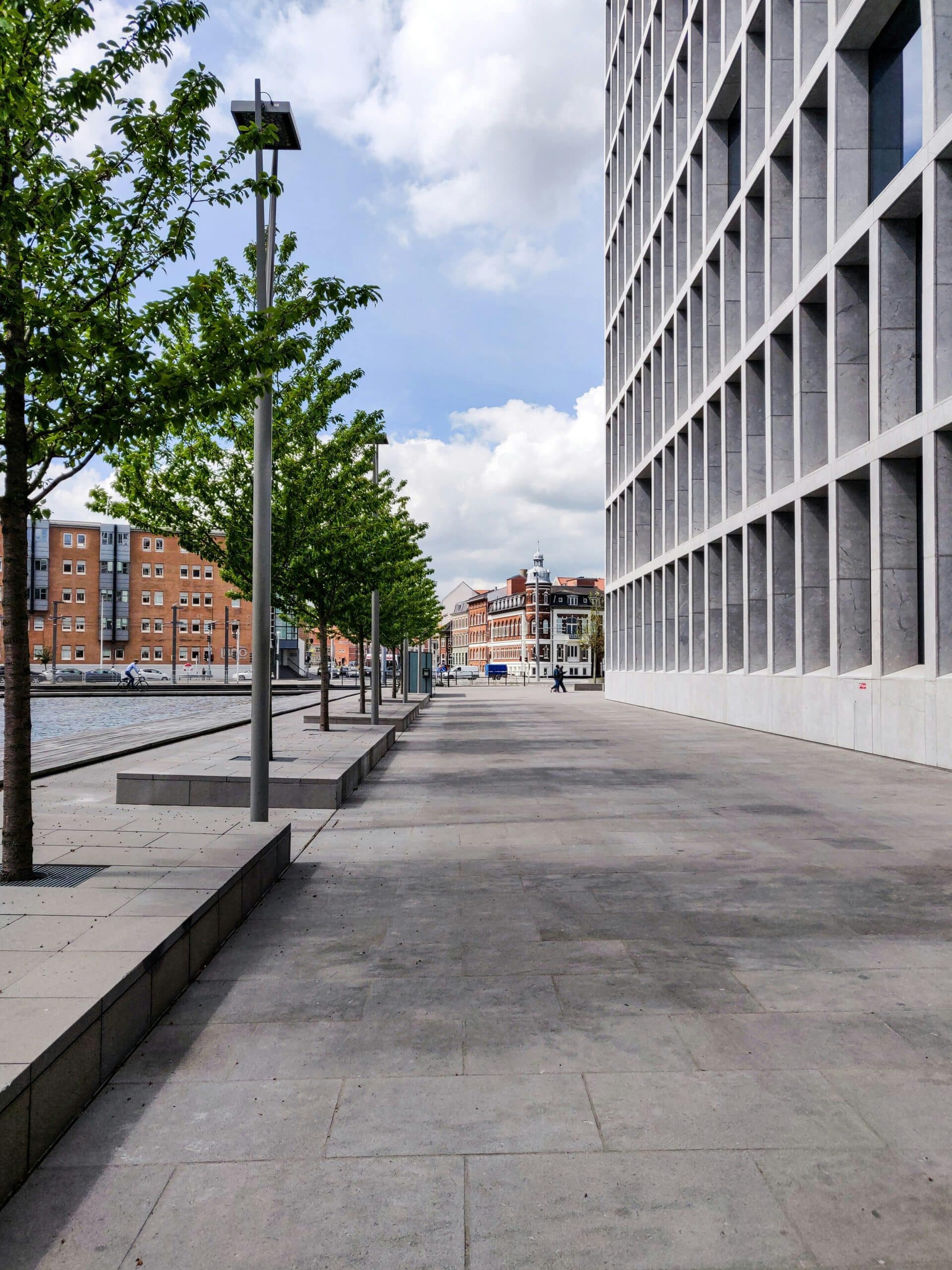 A modern riverside sidewalk, bordered by trees and buildings, including the Frederiksberg Courthouse, with one person visible under a partly cloudy sky.