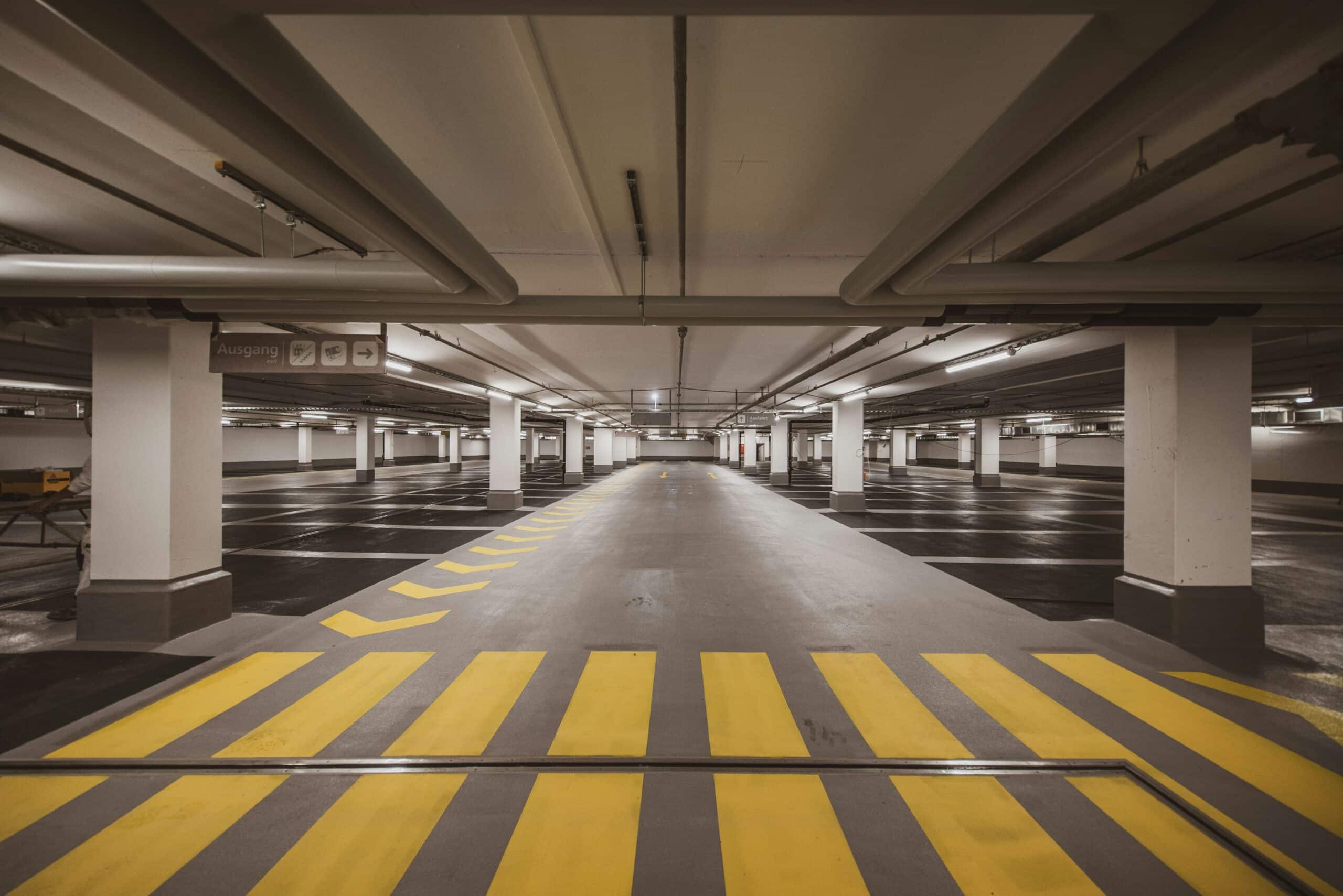 Empty underground parking garage with evenly spaced columns, fluorescent lighting, and yellow-striped pedestrian pathways. Exit signs visible in the distance.