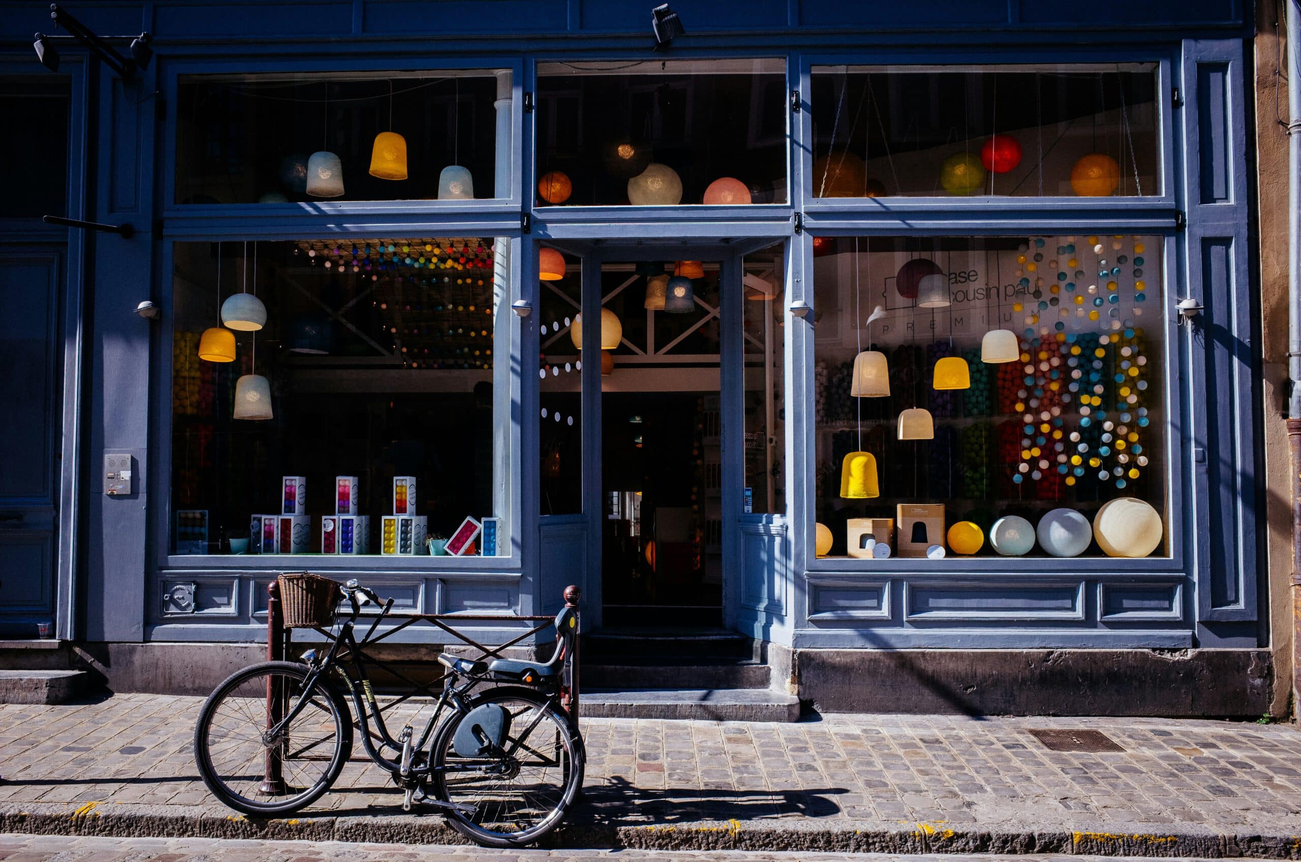 Bicycle parked outside a vibrant light shop with colored lamps displayed in the windows, on a cobblestone street.
