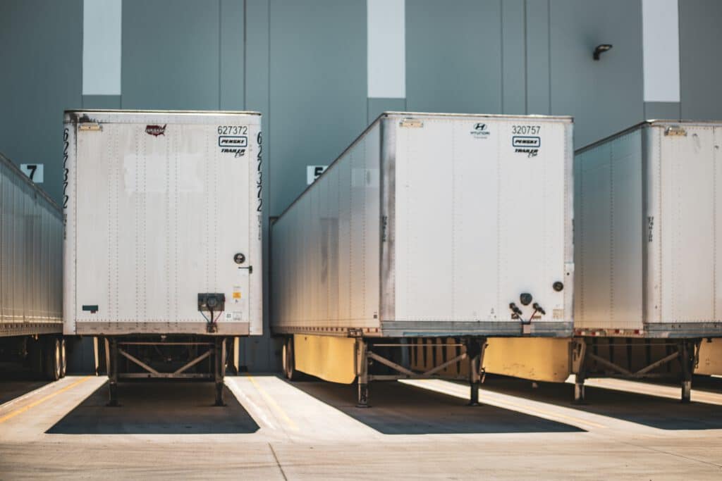 Three parked trailers aligned in an industrial loading area, with numbered dock stations visible in the background, under bright lighting.