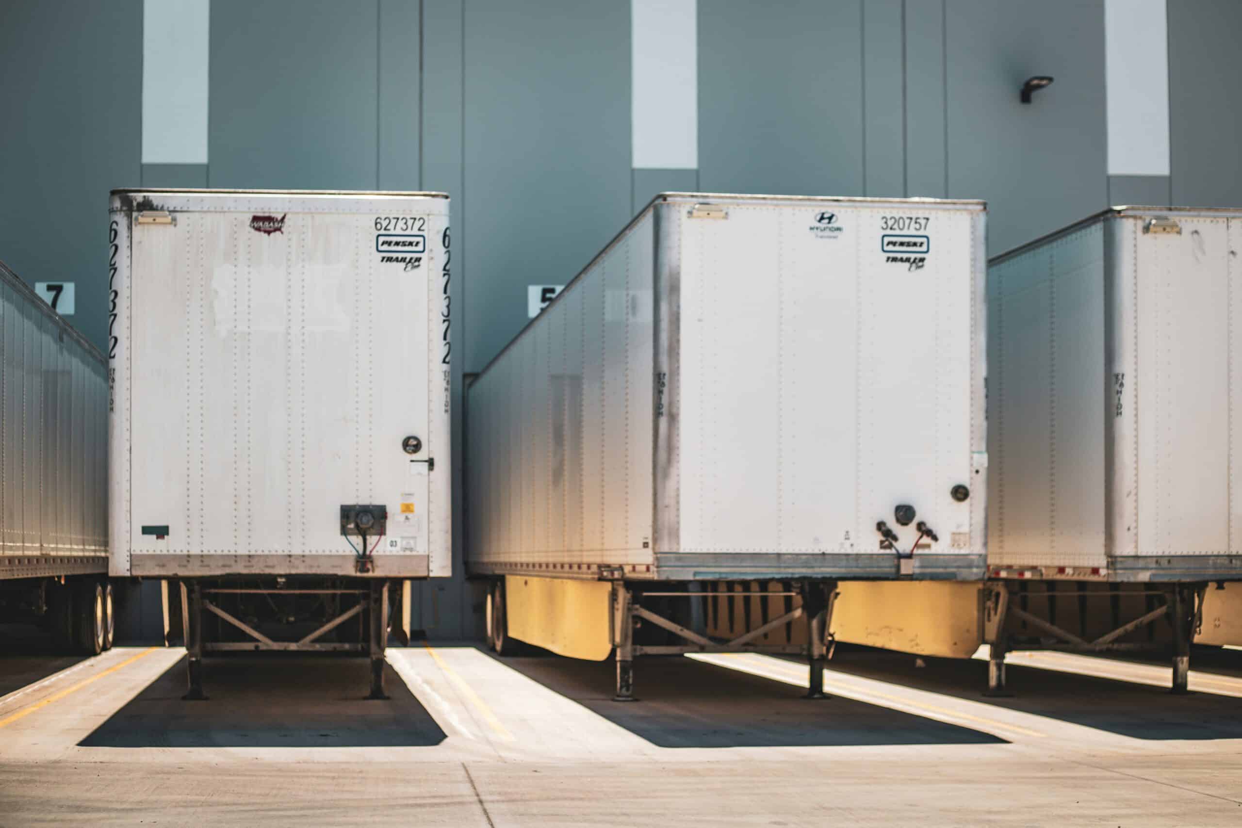 Three parked trailers aligned in an industrial loading area, with numbered dock stations visible in the background, under bright lighting.