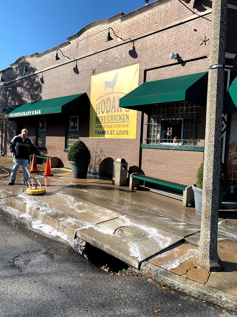 A person pressure washes a sidewalk outside a brick restaurant with a sign thanking St. Louis for voting it best chicken.