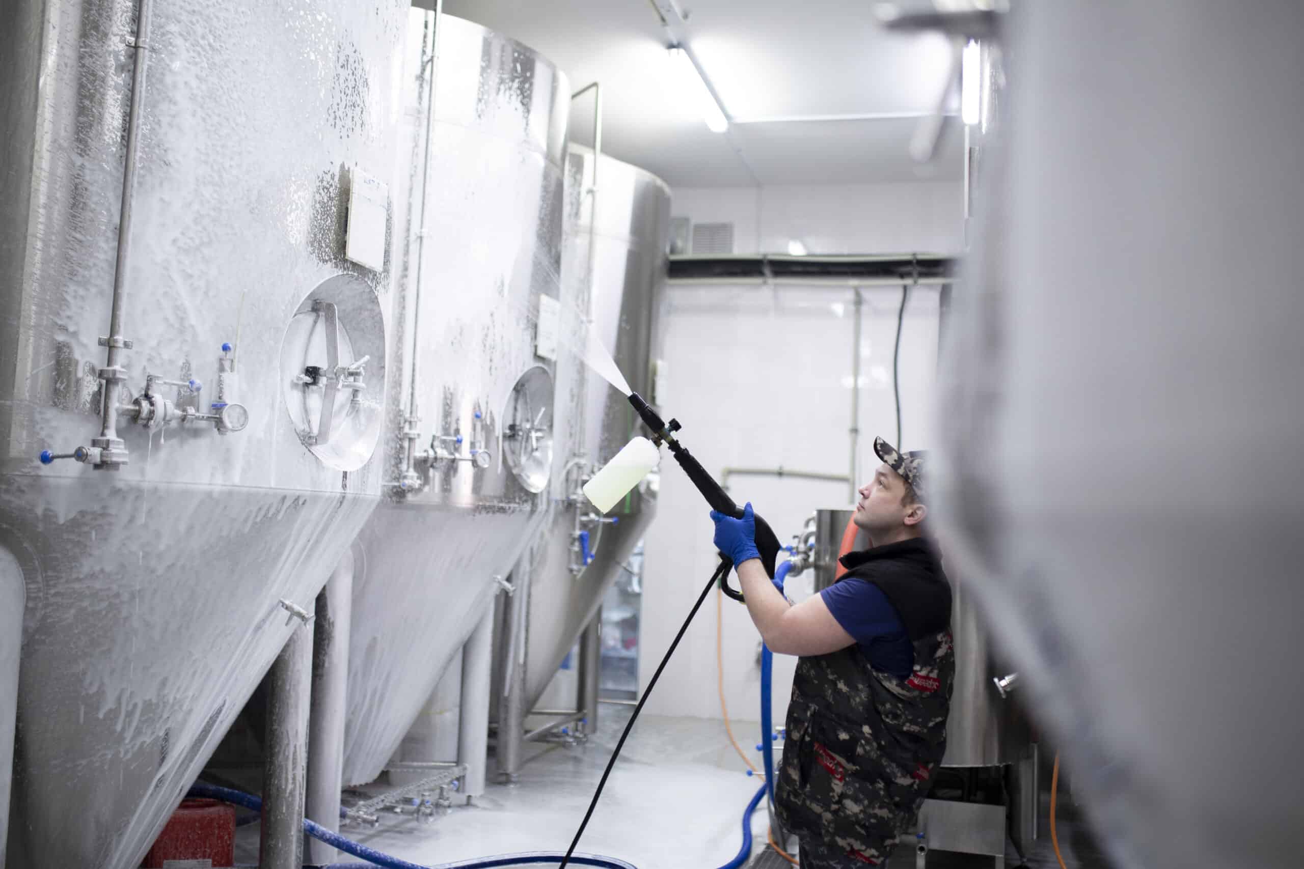 A person in protective gear cleans large industrial tanks with a spray hose inside a modern facility, focusing on hygiene and maintenance.