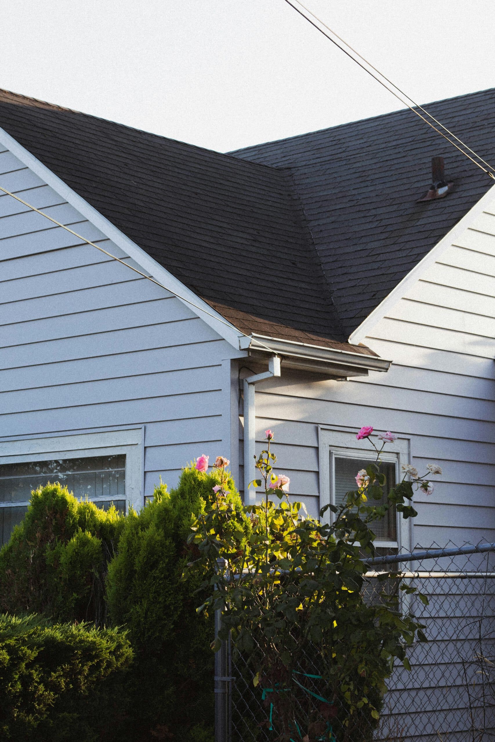 A suburban house with white siding, sloped roof, and vibrant garden seen through a wire fence. Bright flowers add color to the scene.