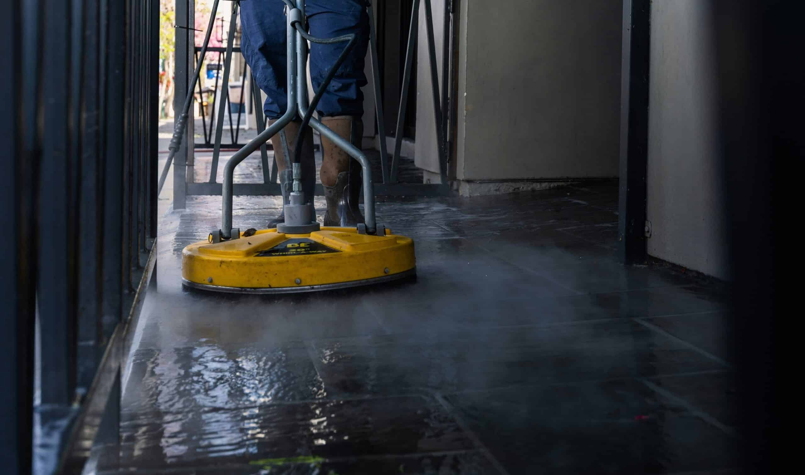 A person cleans a wet, steamy floor using a yellow industrial floor cleaning machine, surrounded by metal railings in a corridor.
